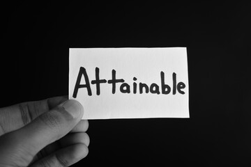 Hand holding a paper card with the word ATTAINABLE