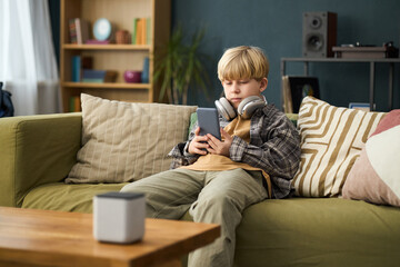 Young boy resting on couch in living room, watching video on tablet while wearing headphones,...