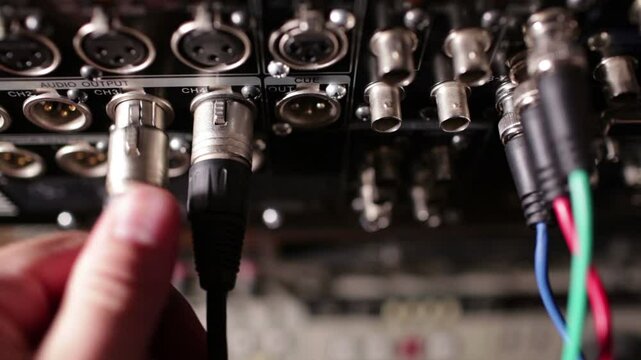 A man is connecting an XLR audio cable to a professional audio output panel labeled "CH3" among various other input/output ports and connectors.
