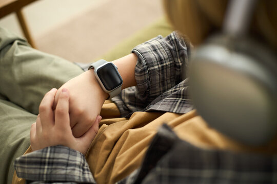 Over-shoulder close-up of anonymous teenager in headphones with smartwatch on wrist resting on couch