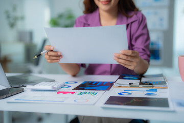 Businesswoman holding and reviewing blank financial report with various charts, graphs, laptop, calculator, and clipboard on white office desk, indicating financial analysis and business planning