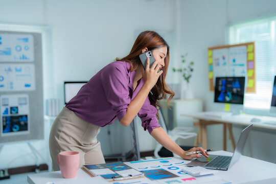Young Asian businesswoman talking on mobile phone and working with laptop computer analyzing financial statistics displayed on desktop and printed charts at office desk