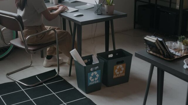 Tilt down shot of female programmer coding on computer and throwing paper into recycling bin while working at desk in cozy office with plants and bicycle by window