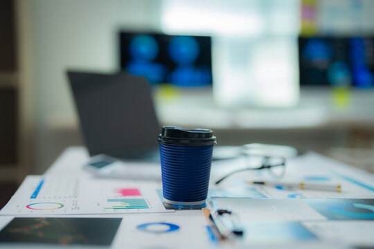 Disposable coffee cup standing on a desk with scattered charts, glasses, pen, calculator and laptop in the background, suggesting a busy work environment during a coffee break