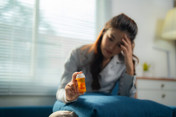 Sad and stressed young woman holding a pill bottle and touching her head while sitting on the sofa...