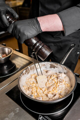 A chef wearing black gloves grinds pepper into a creamy pasta dish with mushrooms in a stainless...