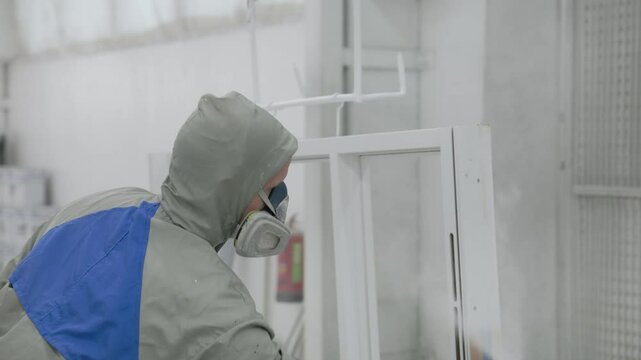 A fully equipped worker in protective gear applies a smooth white coating to a window frame using a spray gun in a clean industrial paint booth environment