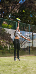 A strong young female athlete suspended in the air mid-leap with her arm extended to return and hit a paddle ball on an outdoor green mountain court.