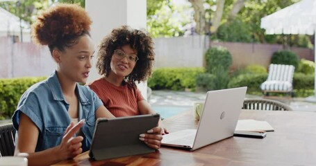 Same-sex lesbian diverse couple embracing joyfully on backyard table with laptop and tablet - Powered by Adobe