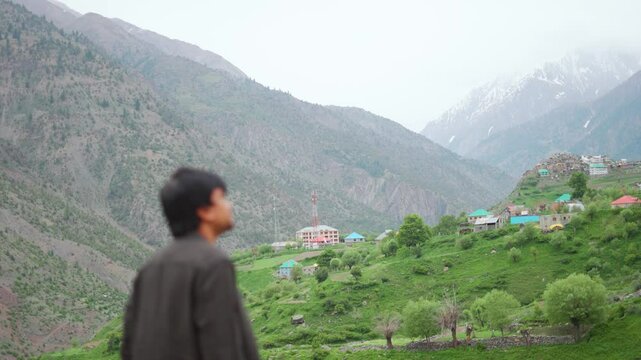4K shot of a defocused Indian man looking at Triloknath village in front of the snowy Himalayan mountain peaks at Pattan valley in Lahaul, Himachal Pradesh, India. Man looking at the village on cliff.