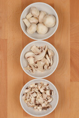 Three bowls with mushrooms, ranging from whole to finely chopped, on a wooden surface, shot from above