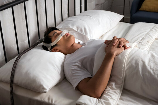 Asian man sleeping calmly with CPAP nasal mask in bed, a treatment for snoring and obstructive sleep apnea (OSA). Promotes better breathing and restful sleep with home respiratory therapy.