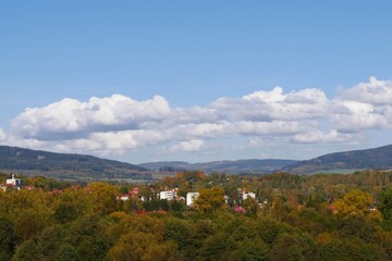 Panoramic view of a town nestled amongst colorful autumn trees, with rolling hills in the background under a blue sky with white clouds - Kamienna Góra, Poland