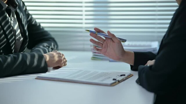 Two people are sitting at a table with a clipboard in front of them. One of them is holding a pen and pointing at the clipboard. Scene is professional and serious