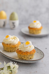 Lemon Poppy Seed Cupcakes with lemon curd and cream cheese topping and Fried Egg candies. Colourful Easter eggs, flowers on a stone background, top view. Easter holiday and festive food.