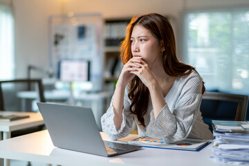 A woman is sitting at a desk with a laptop and a stack of papers