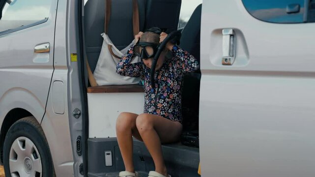 Young girl wearing a swimsuit gets ready to snorkel, holding her mask while sitting on a van step