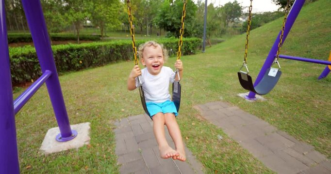 Cheerful tiny boy wearing white t shirt and blue shorts, happily swinging on swing set in playground, slow motion shot. Dynamic wide angle camera move forth and back to keep emotional kid in frame