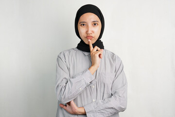Fototapeta premium Confused Southeast Asian woman in headscarf and shirt, thinking pose with hand on chin. Studio portrait on white background, faceless expression concept, minimalist photo.