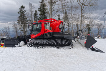 A red snow groomer sits atop a snowy peak, ready to smooth the slopes, surrounded by bare trees under a cloudy, overcast sky.