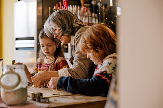 A woman is baking cookies with a young girl and a red-haired boy in a cozy kitchen. The children are focused on the dough as they cook together during a holiday at home.