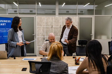 In an office, colleagues hold a business meeting around a conference table. An Asian woman presents near a screen, while an older White man talks with a teammate. Concept of teamwork and discussion.