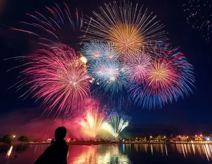 Fireworks bursting over a summer festival at night, reflected on water with a silhouette viewer in the foreground 