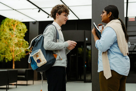 A White young man with glasses, wearing a gray sweater and carrying a backpack, talking with an Indian young woman in professional attire, both having a conversation during an internship.