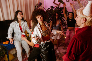 A multiethnic group of young men and women is dancing, singing, and holding drinks at a lively house party with music and decorations in a cozy indoor setting.