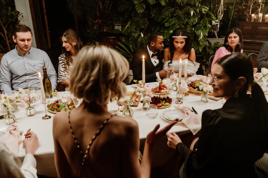 A Black young bride and groom seated closely at a candlelit wedding reception table surrounded by a multinational group of wedding guests enjoying a festive evening meal in an elegant indoor venue.