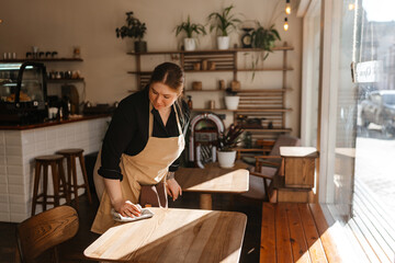 A White adult woman wearing an apron wipes down a wooden table inside a modern cafe during the day. Sunlight streams through the large front window as she prepares the interior space for customers. © Drobot Dean