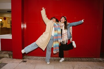 Asian and Hispanic adult women wearing scarves and winter coats pose joyfully in front of a bright...