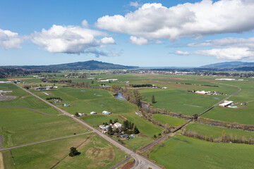 Fototapeta premium Aerial views of lush green countryside in Tillamook, Oregon, a small coastal town near the Pacific Ocean, showing farmlands, country roads, rural houses, mountains, and blue sky in spring-summer