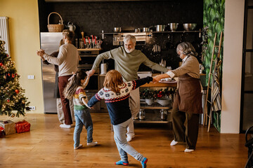 A smiling White family, including grandparents with their two grandchildren who hold hands and dance, and their parents dancing next to them, in a decorated kitchen near a decorated Christmas tree.