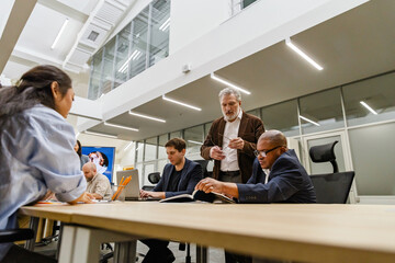 A 55-year-old White businessman dressed in a brown business suit, standing next to the table of his colleagues in an office room, as they're having a meeting, and reviewing some papers of his coworker