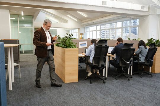 A mid-50s Caucasian businessman in glasses, reviewing his clipboard with a pencil in his hand as he walks around the spacious office hall, with his coworkers sitting at their cubicles
