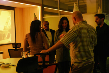 A mid-30s Asian woman wearing a business suit, standing among a diverse group of colleagues in a dimly-lit office hall, lighting the candles on a cake as they're having an after-work party