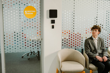 A young White man aged 25, with curly brown hair and glasses, wearing a gray business suit, sitting...