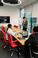 Diverse colleagues gather around a conference table in a modern office. A young White man stands and presents to colleagues. The room contains large windows, a TV screen, and colorful chairs.