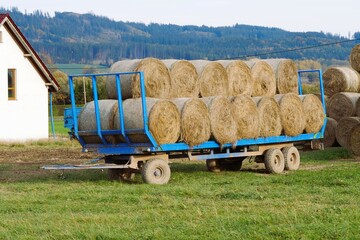 Two-axle trailer loaded with round hay bales sits in a grassy field near a white building, with hills and trees in the background