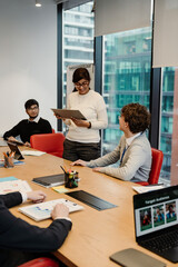 A smiling brunette Indian woman in her 30s stands at a table where her multiethnic colleagues sit and look at her as she reads something from a clipboard, in a meeting room, with a large window.