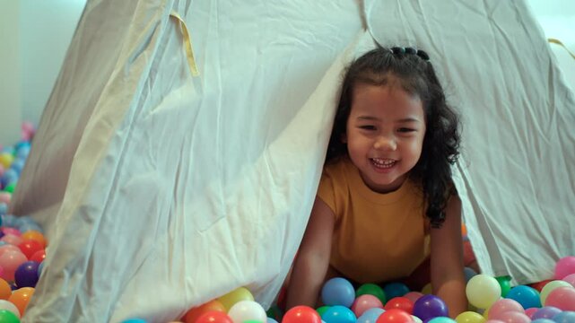 Happy little girl plays hide and seek inside teepee tent surrounded by colorful ball pit, promoting fun. Child development through imaginative indoor play.
