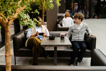 Obraz premium An Indian woman in her 20s with glasses, and a blue shirt, and a White man in his 20s with glasses and a grey cardigan sit on a black couch at a table in a modern office lobby having a lunch break.
