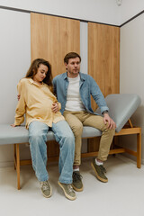 Obraz premium A lovely White couple in their mid-30s, a pregnant wife and her husband, sitting on a medical bed in a white doctor's office, waiting for something as the wife caresses her stomach
