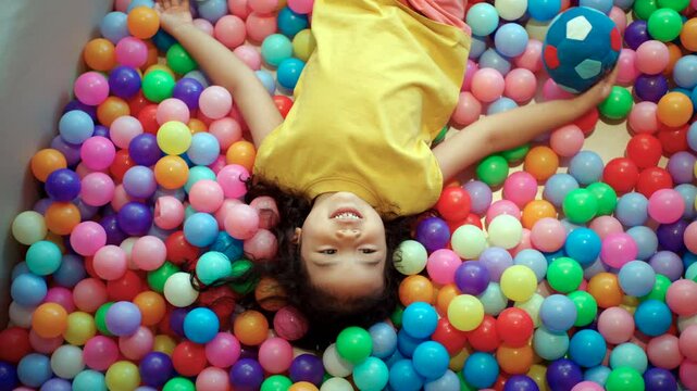 A happy little girl plays with a ball in a colorful ball pit, enjoying the fun and enhancing her development through playful activities and exercise.
