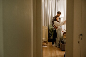 A view from a hallway of a smiling White couple in their 30s in home clothes standing near a bed in...