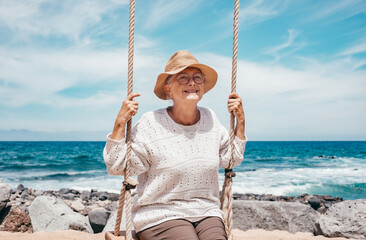 Happy senior woman sitting on swing on sea beach enjoying relaxed and carefree moments, positive...