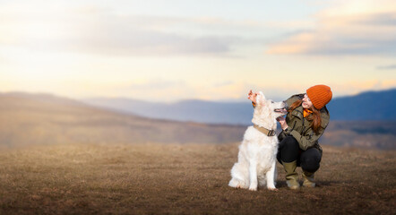 A cheerful young woman in outdoor clothing next to her golden retriever dog on a mountain meadow during sunset. Travel with dog. Life with dog. © kathrineva20