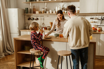 A loving European family of three, standing around the table in their modern kitchen, as the mid-30s parents make a breakfast for their school-aged son, who sits beside them, dressed in pajamas