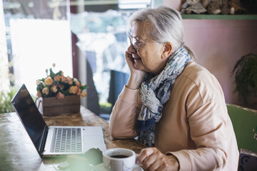 White haired senior woman wearing eyeglasses sitting indoor using laptop while has a coffee break, addicted  elderly lady enjoying food and drink